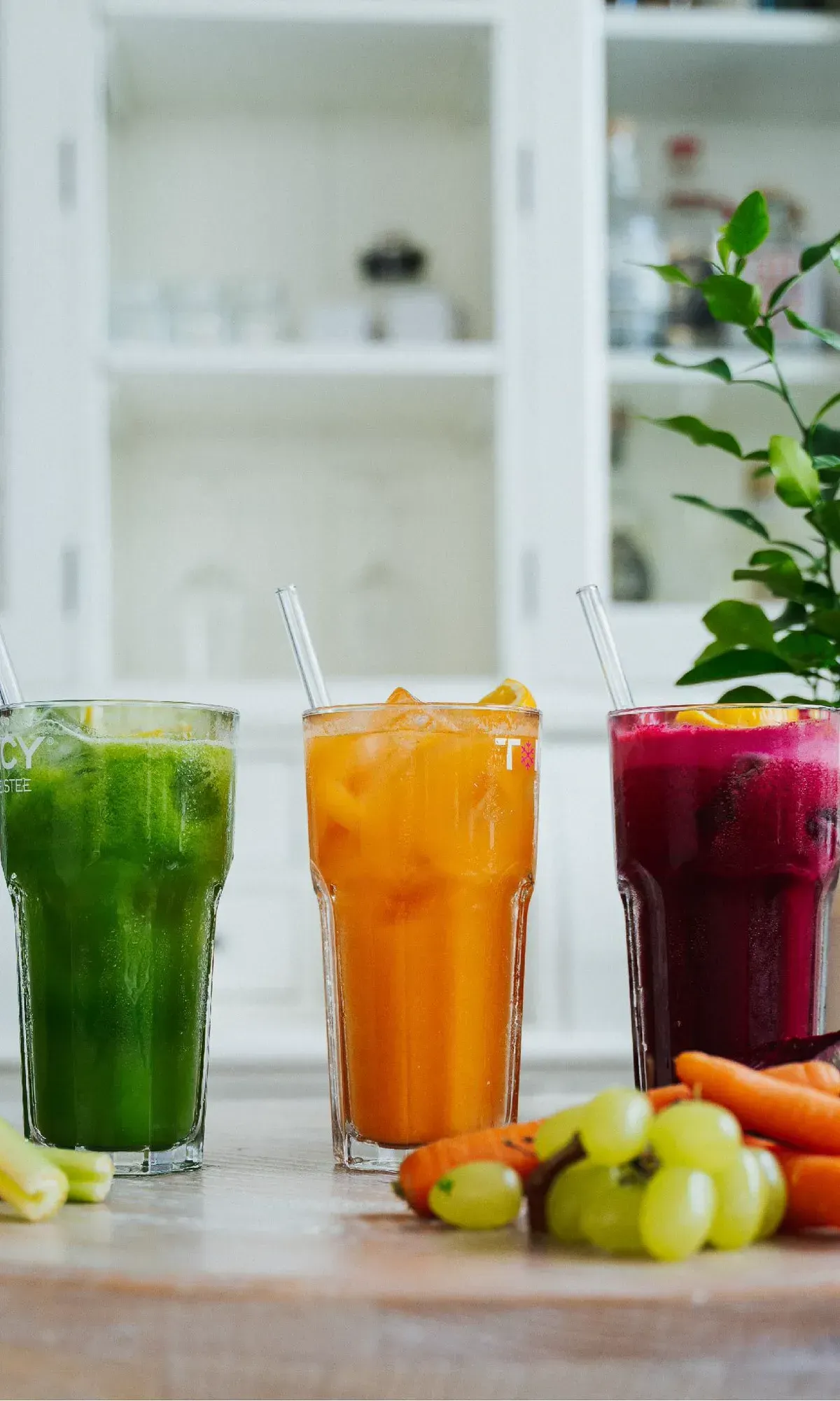 Drei hohe Gläser mit frisch gepressten Säften in Grün, Orange und Beere, daneben Obst und Gemüse.